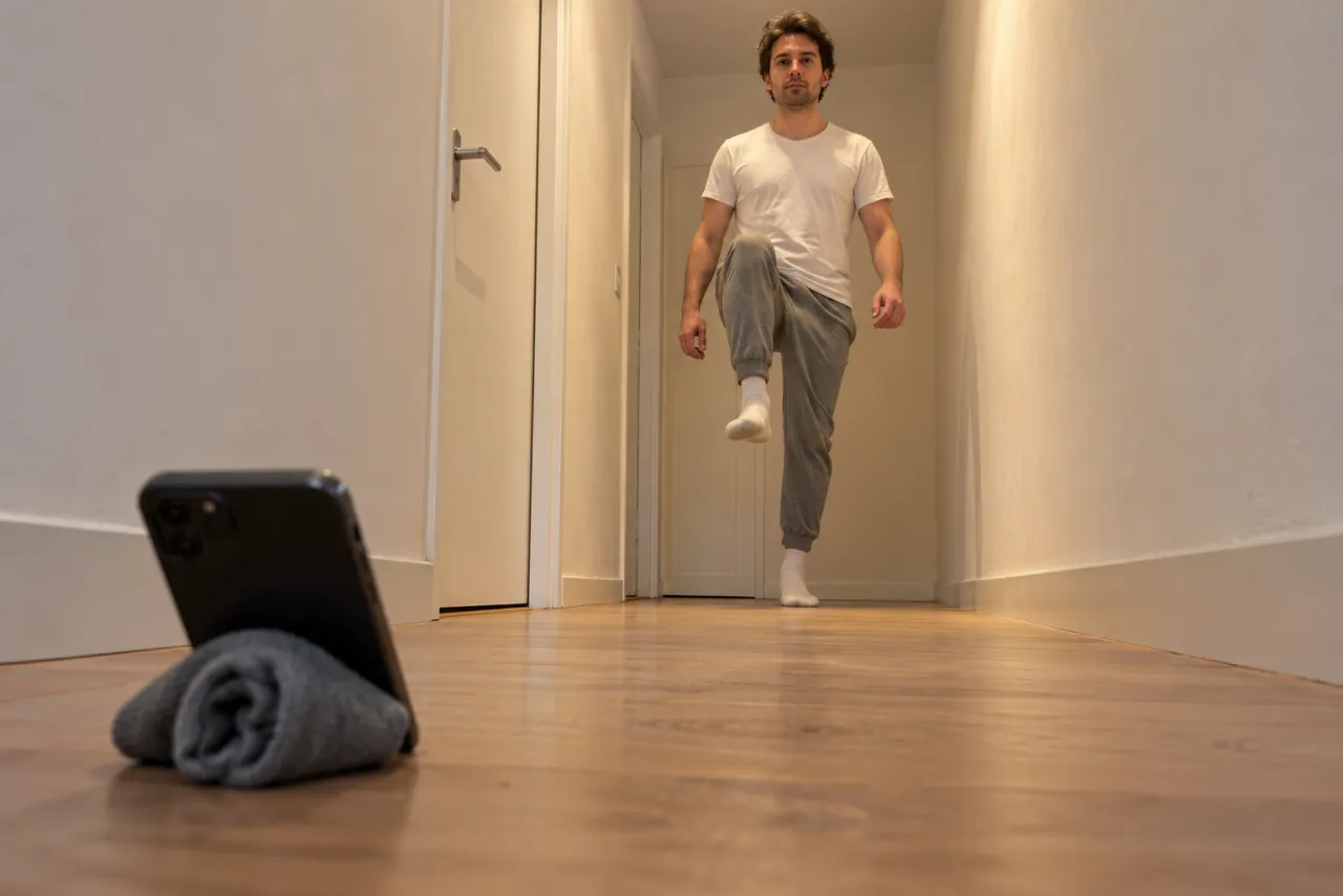 Low angle from behind a phone propped on a rolled grey sock on a wooden hallway floor, as a man in a white tee and grey joggers marches toward it with one knee high