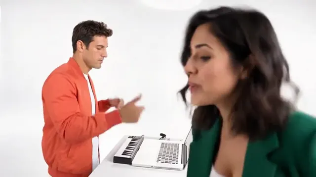 A man in an orange jacket plays a mini keyboard while a woman in a green jacket leans into the lens