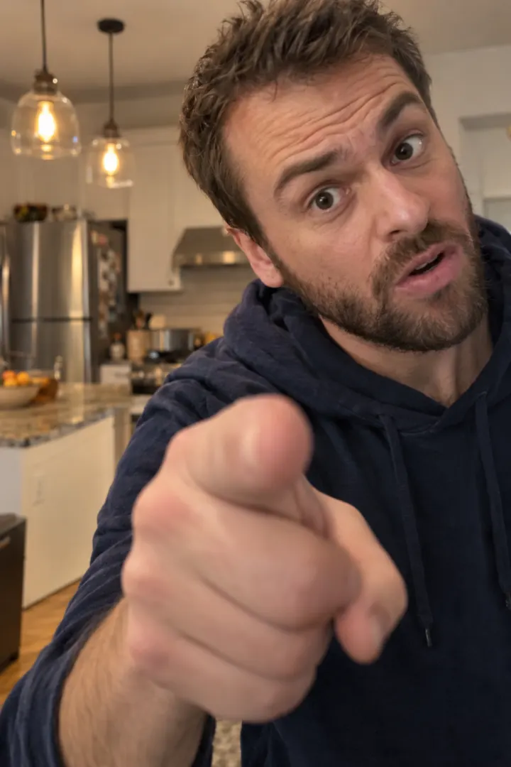 A man in a navy hoodie points straight at the camera in a kitchen, mid-rant