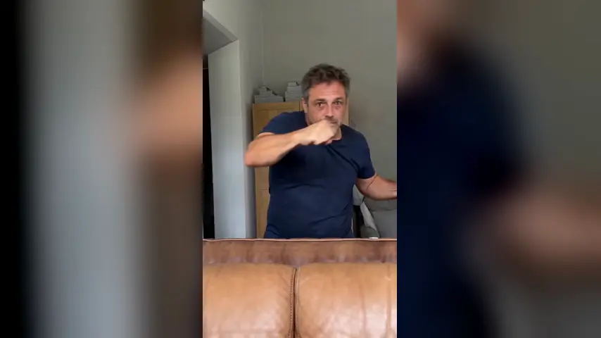 A young man in a navy t-shirt throws a hook behind a brown leather sofa in his living room, a phone propped on the seat