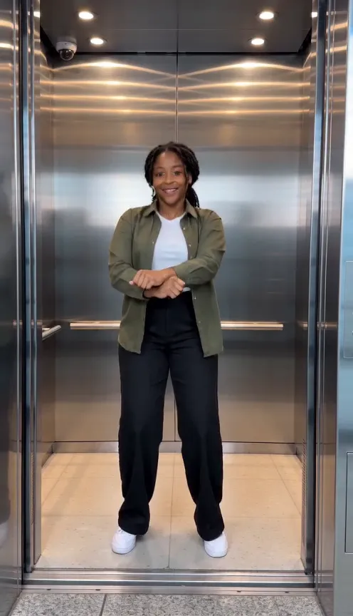 A smiling woman in an olive overshirt, white tee and black wide-leg trousers does the crossed-wrist trot inside a lit steel elevator with its doors wide open