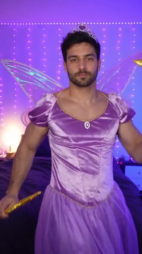 A bearded dark-haired man in a lilac satin fairy dress with wings and a tiara holds a star wand in a purple bedroom with fairy lights