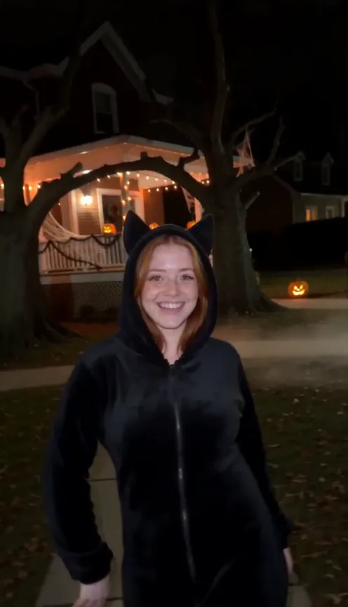 A woman in a black hooded onesie with pointed cat ears smiles on a street with string lights